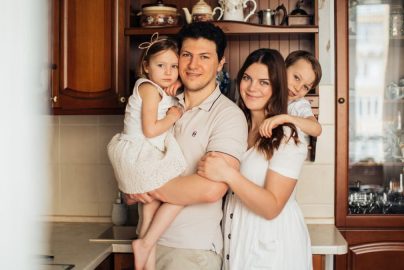 Satisfied man with daughter and woman with son embracing embracing each other while standing against wooden cupboard in kitchen and looking at camera