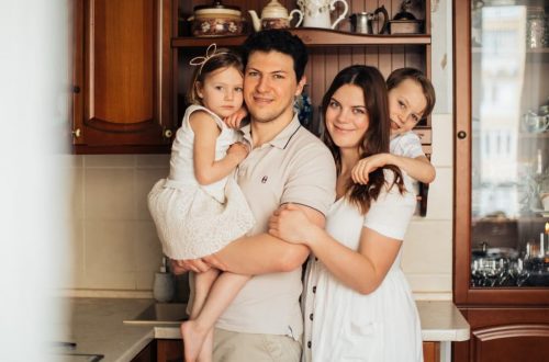 Satisfied man with daughter and woman with son embracing embracing each other while standing against wooden cupboard in kitchen and looking at camera