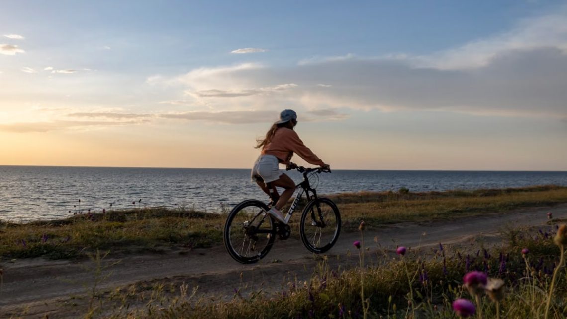 A woman rides her bicycle along a seaside path under a serene sunset, embracing nature.
