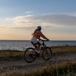 A woman rides her bicycle along a seaside path under a serene sunset, embracing nature.