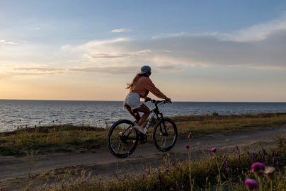 A woman rides her bicycle along a seaside path under a serene sunset, embracing nature.