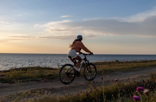 A woman rides her bicycle along a seaside path under a serene sunset, embracing nature.